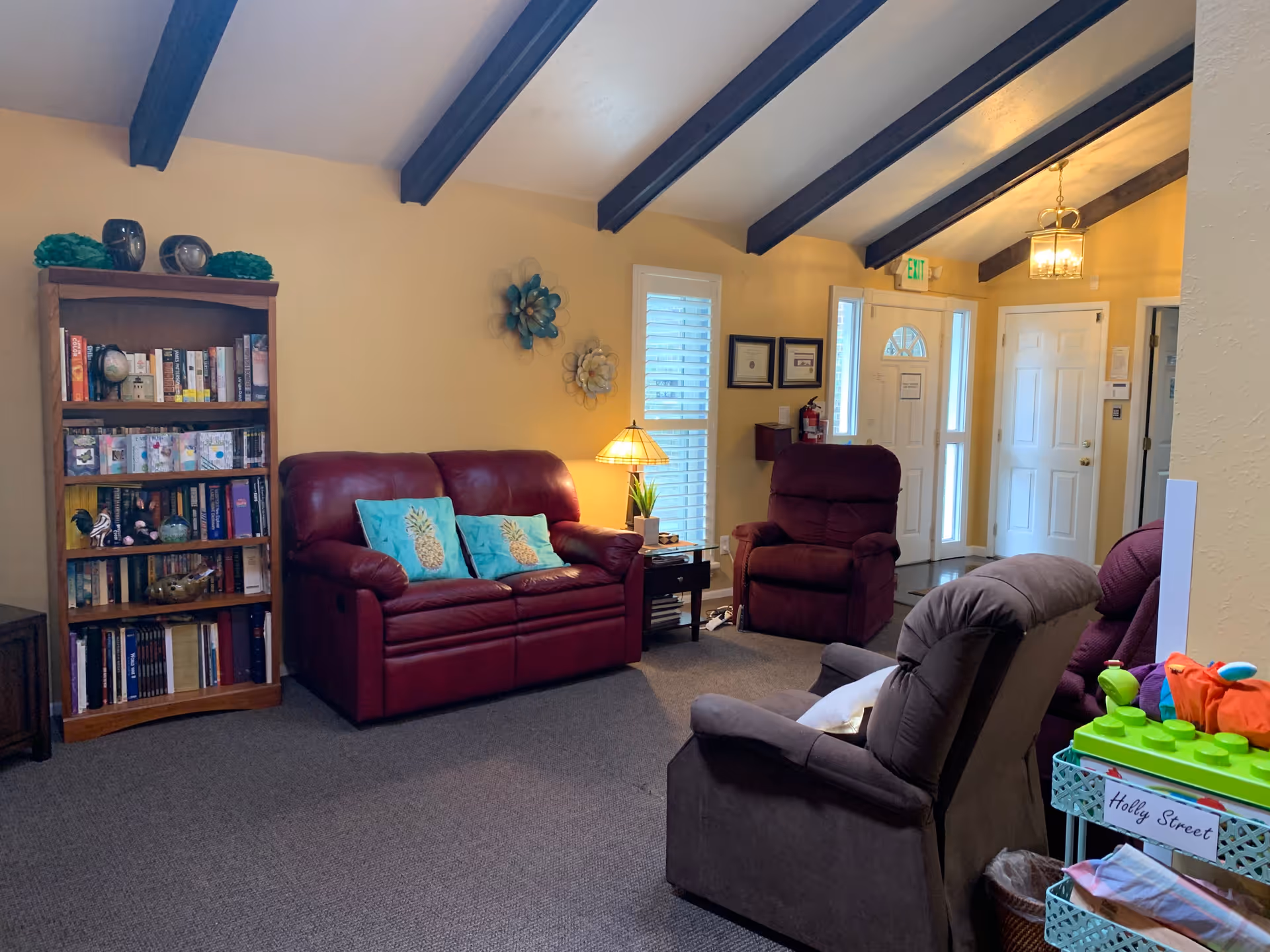 A cozy living room with a burgundy leather loveseat adorned with two turquoise pineapple pillows, a matching burgundy recliner, and a gray recliner chair. There is a wooden bookshelf filled with books and decorative items on the left side. A small side table with a lamp and a plant is positioned between the loveseat and the recliner. The room has yellow walls, exposed dark wooden ceiling beams, and a carpeted floor. Near the entrance door, there are framed certificates on the wall and a fire extinguisher. A small cart labeled 'Holly Street' with toys and blankets is visible on the right side.