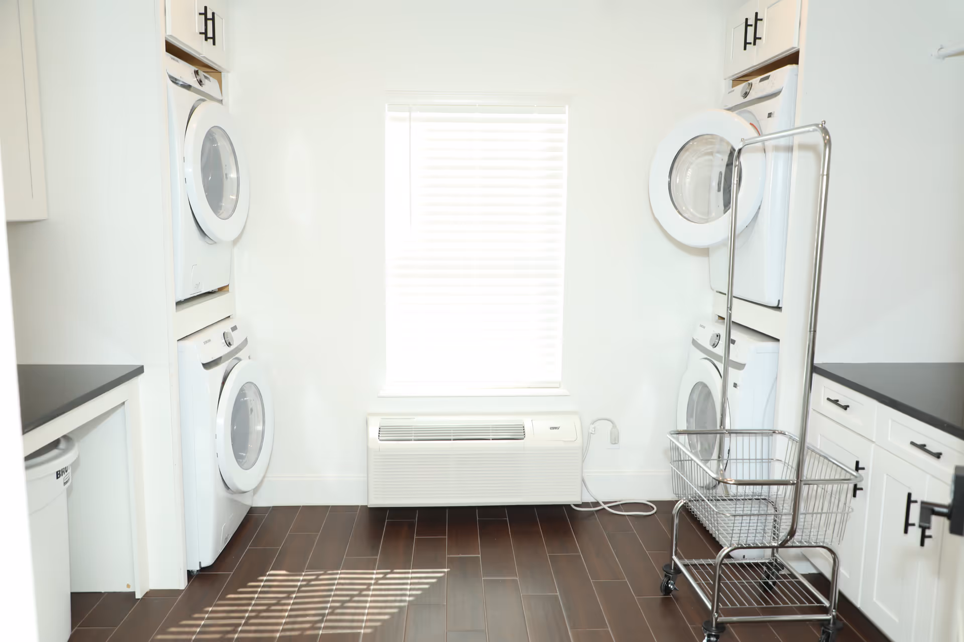 Bright laundry room with two stacked washer and dryer units on each side, white cabinetry with black handles, a metal laundry cart with wheels, a window with white blinds, and dark wood flooring.
