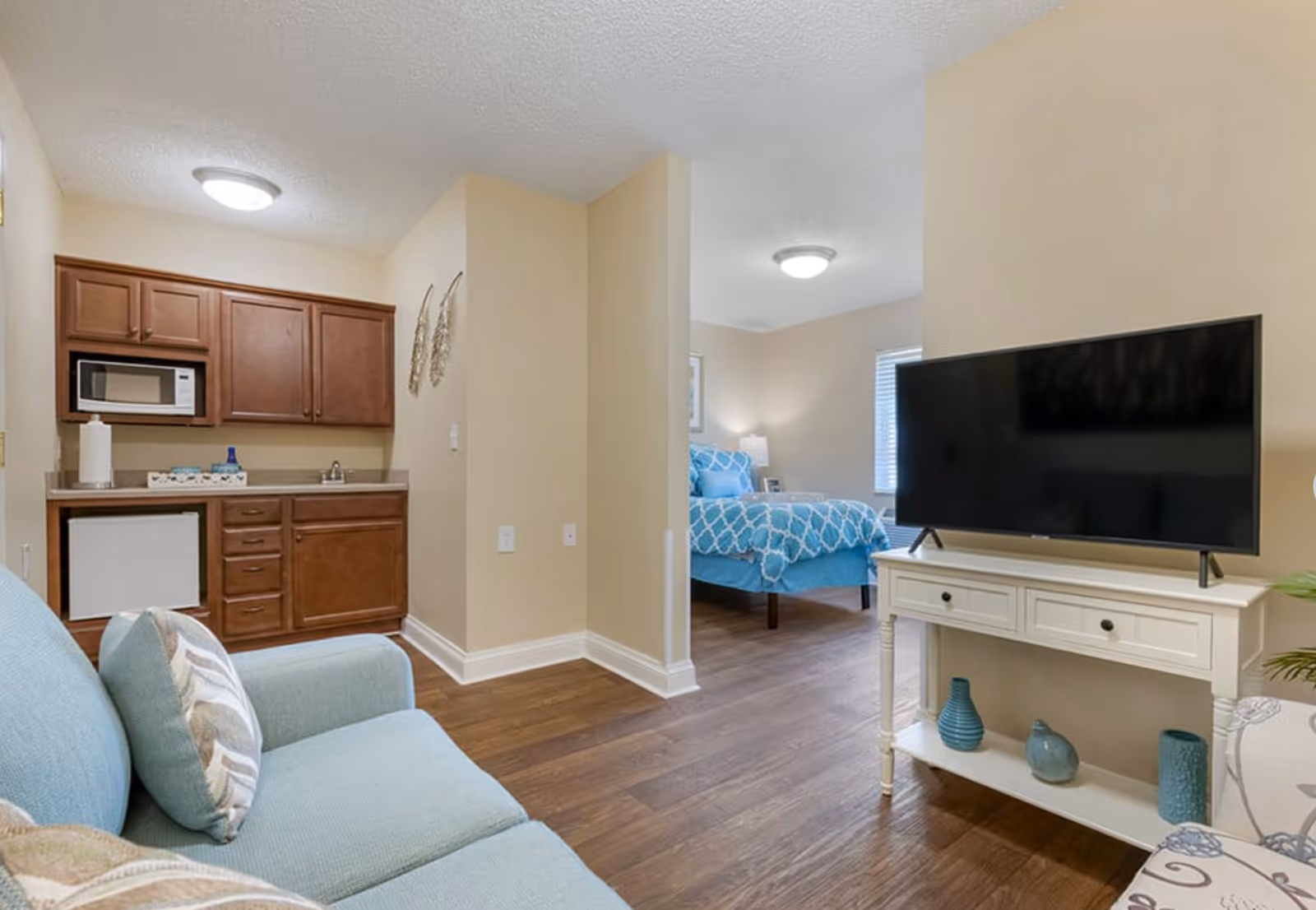 Studio apartment interior featuring a kitchenette, a teal sofa, a TV on a white console, and a bed in the background.