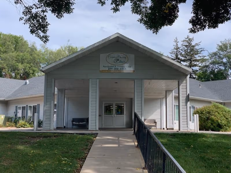 Front exterior view of Corridor Crossing Place, a single-story building with a covered entrance supported by columns. There is a concrete walkway with a handrail leading up to double glass doors. The building is surrounded by green grass and trees.