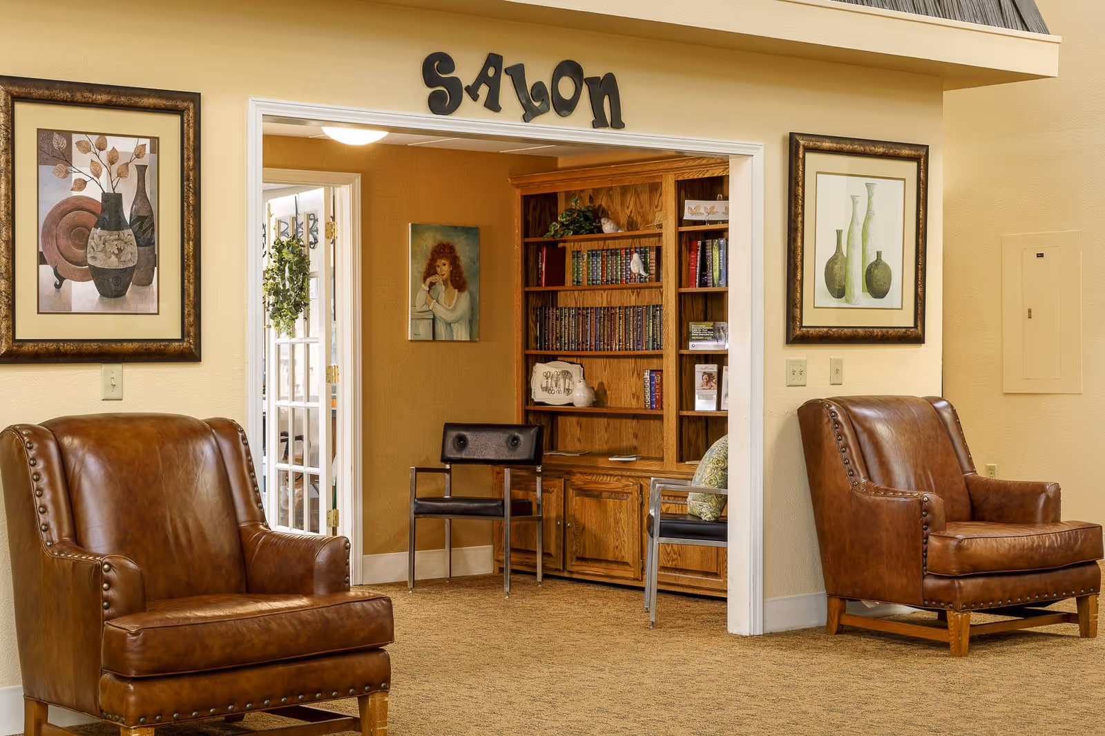 A cozy assisted-living common area with two brown leather armchairs flanking a doorway labeled "SALON" that opens to a bookshelf and seating.