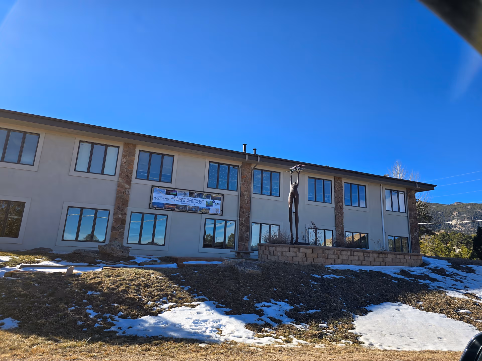 Exterior view of a two-story building with multiple windows, a stone and stucco facade, and a tall abstract metal sculpture in front. There is some snow on the ground and a clear blue sky above.