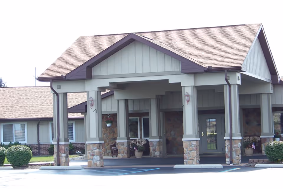 Front entrance of a single-story care building with a covered porte-cochère supported by stone-based columns, potted plants, and a glass door.