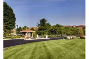 Well-maintained front lawn and driveway with a small gazebo-like entrance and apartment buildings in the background under a clear blue sky.