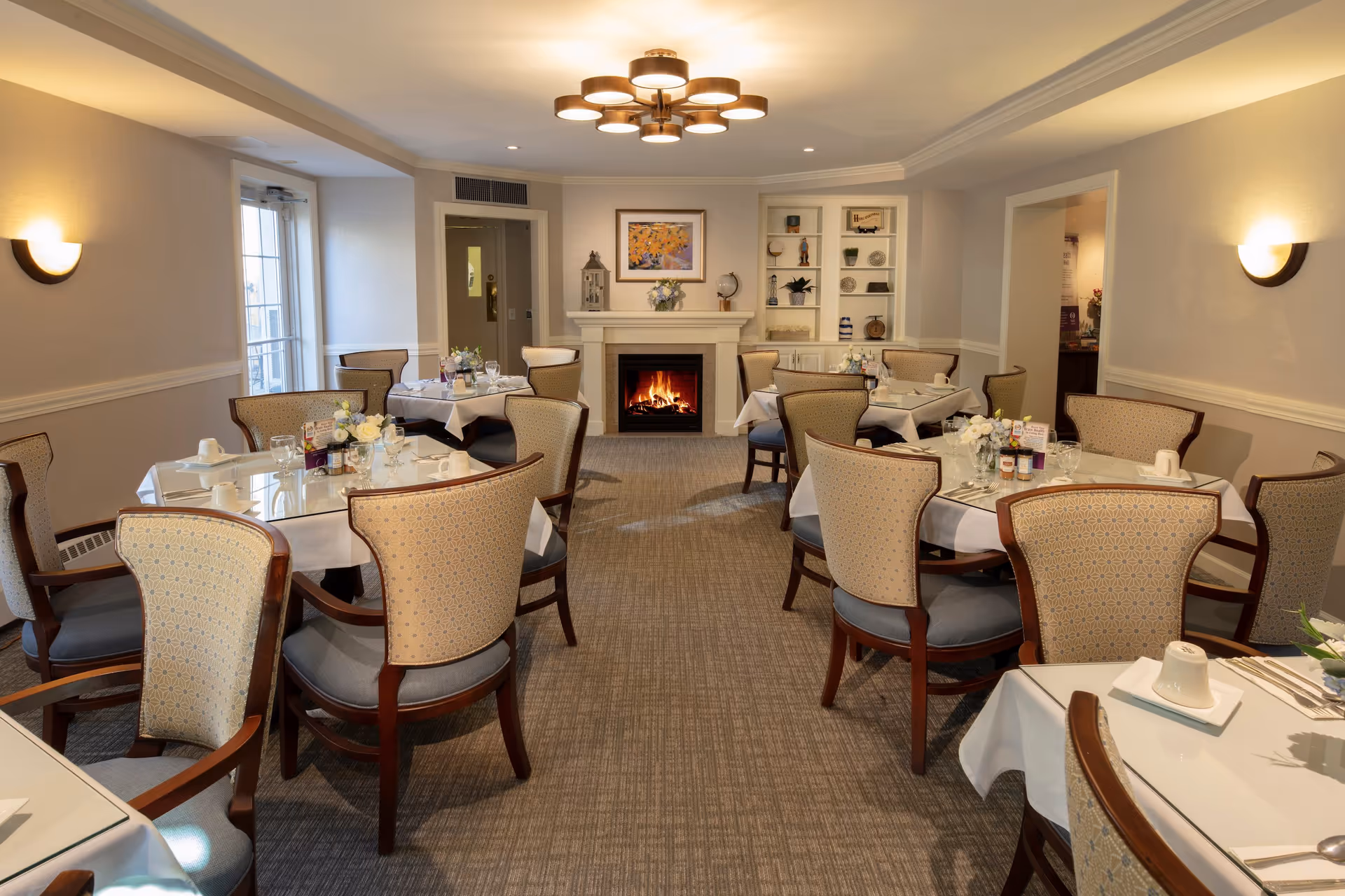 A cozy dining room with multiple tables covered in white tablecloths, each set with cups, glasses, and small flower arrangements. The room features upholstered chairs with wooden frames, a lit fireplace at the far end, built-in shelves with decorative items, and soft wall lighting. A modern ceiling light fixture hangs above, and there is a door leading outside on the left side.