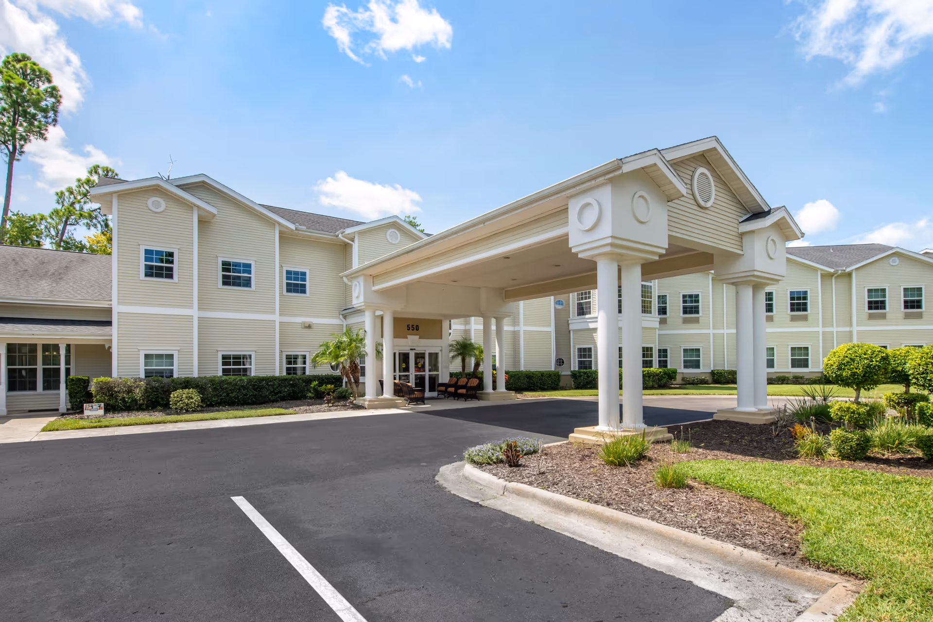 Exterior view of Brookdale Ormond Beach senior living facility showing a large covered entrance with white columns, beige siding, multiple windows, landscaped greenery, and a clear blue sky.