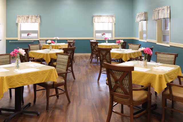 A dining room with several tables covered in yellow tablecloths, each set with white napkins and small vases holding pink flowers. The room has wooden floors, light blue walls with a cream-colored chair rail, and windows with white curtains letting in natural light.