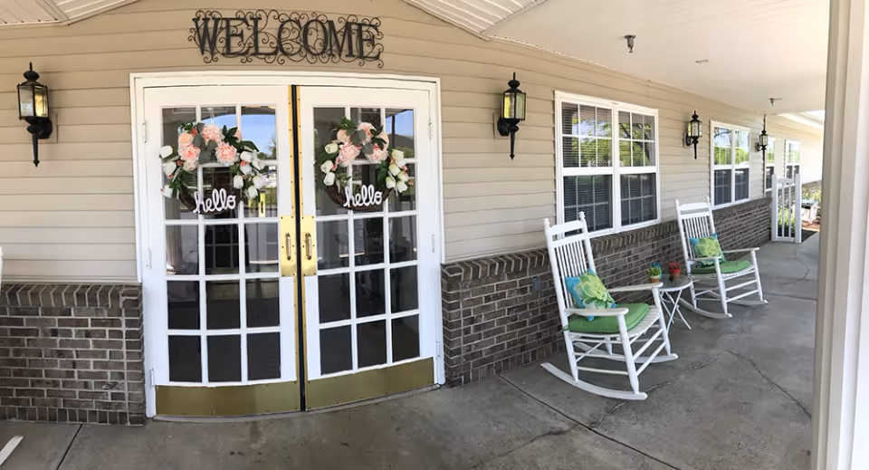 Entrance to a facility with double glass doors decorated with floral wreaths that say 'hello'. Above the doors is a metal sign that reads 'WELCOME'. To the right of the doors, there are two white rocking chairs with green cushions and small side tables with potted plants. The exterior wall is beige with brick accents and several windows with white frames.