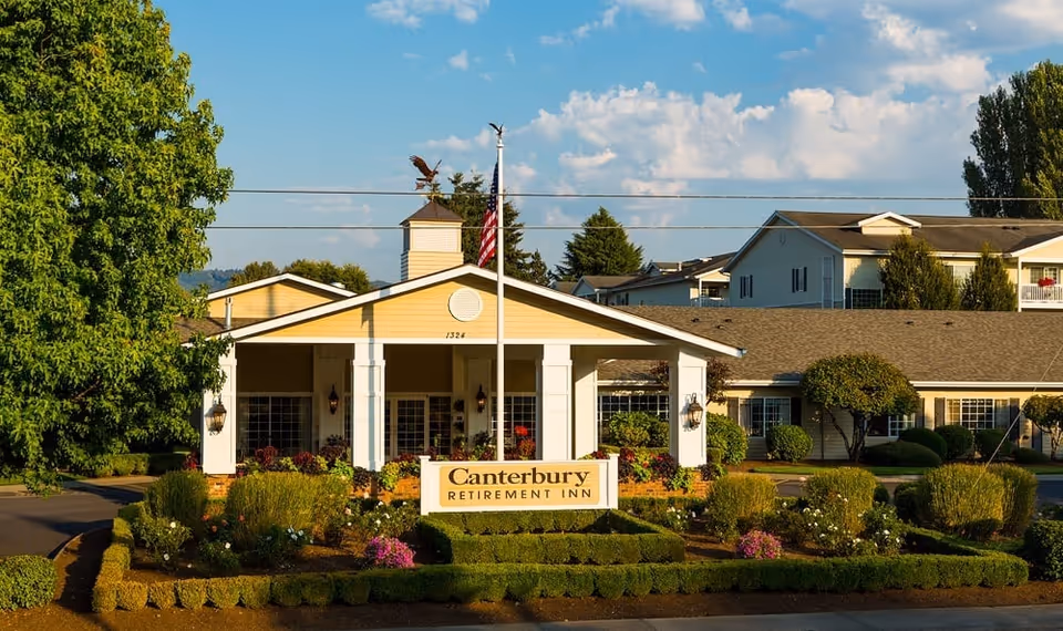Front exterior view of Canterbury Retirement Inn building with a well-maintained garden and American flag in front under a partly cloudy sky.