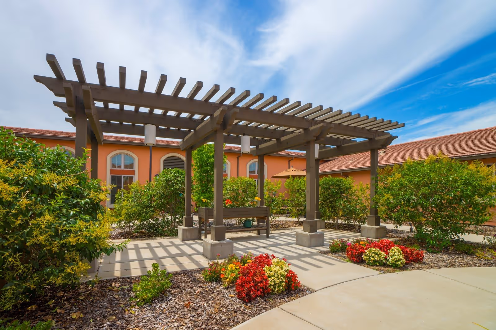 Outdoor garden area at Eskaton Village Roseville featuring a wooden pergola with hanging lights, surrounded by green bushes and colorful flowers, with a building with arched windows and a red-tiled roof in the background under a partly cloudy blue sky.