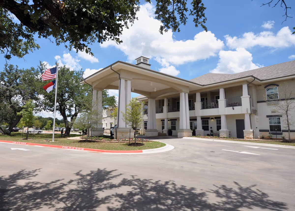 Exterior view of The Legacy at Falcon Point senior living facility showing a two-story building with a covered entrance supported by large columns, surrounded by trees and a driveway. Two flagpoles display the American flag and another flag under a partly cloudy blue sky.