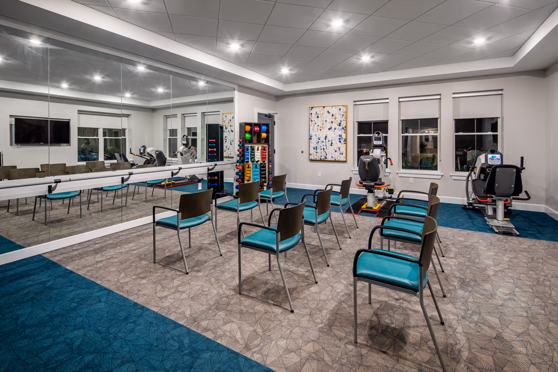 A spacious fitness/activity room with teal chairs arranged in rows, exercise equipment, a wall of mirrors and a rack of colorful weights.
