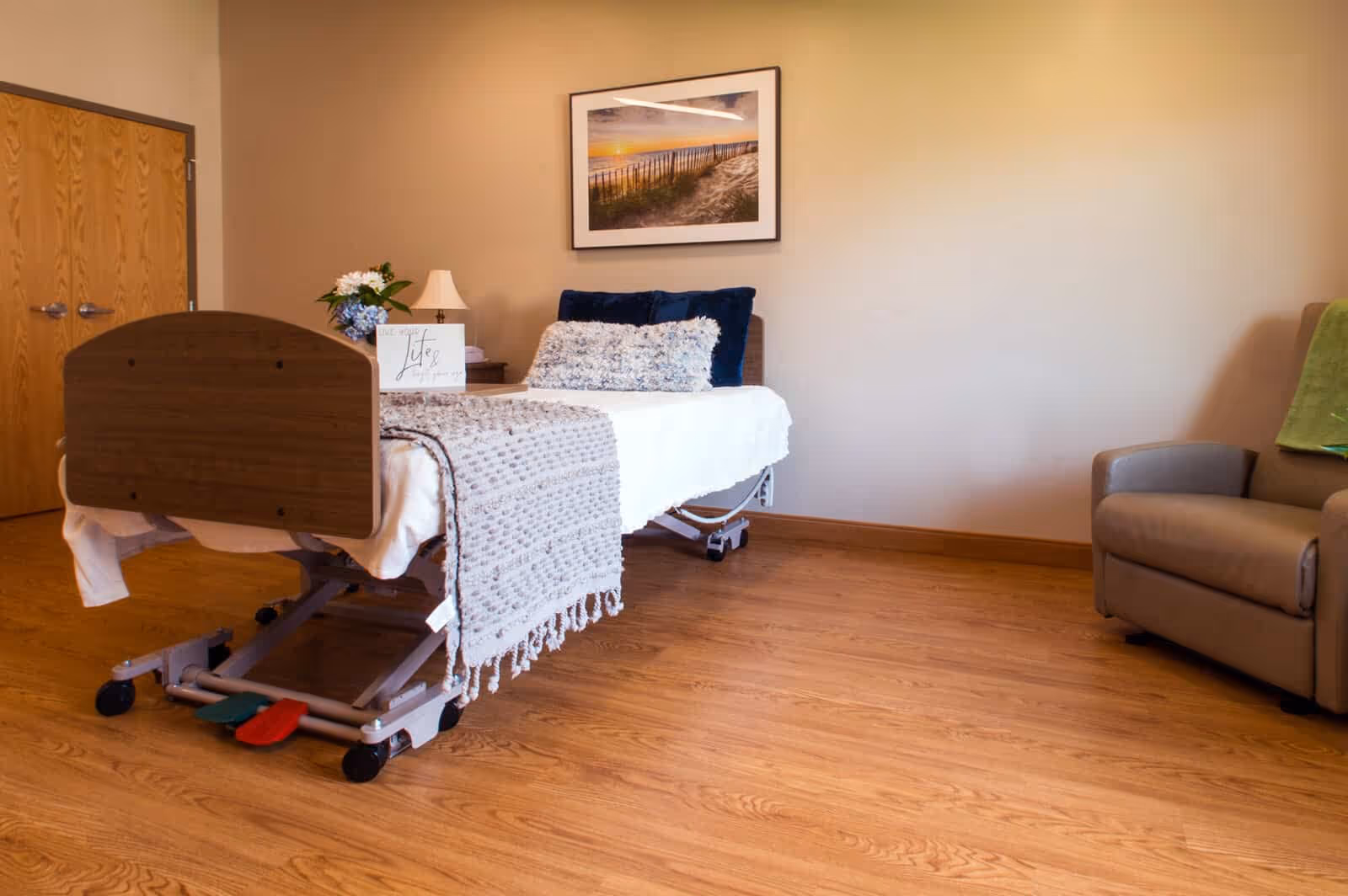 A neatly arranged healthcare facility bedroom with a hospital bed covered in white sheets, a textured blanket, and multiple pillows. There is a wooden door on the left, a nightstand with a lamp and flowers, a framed picture of a beach scene on the wall, and a comfortable armchair with a green blanket on the right. The room has wooden flooring and beige walls.