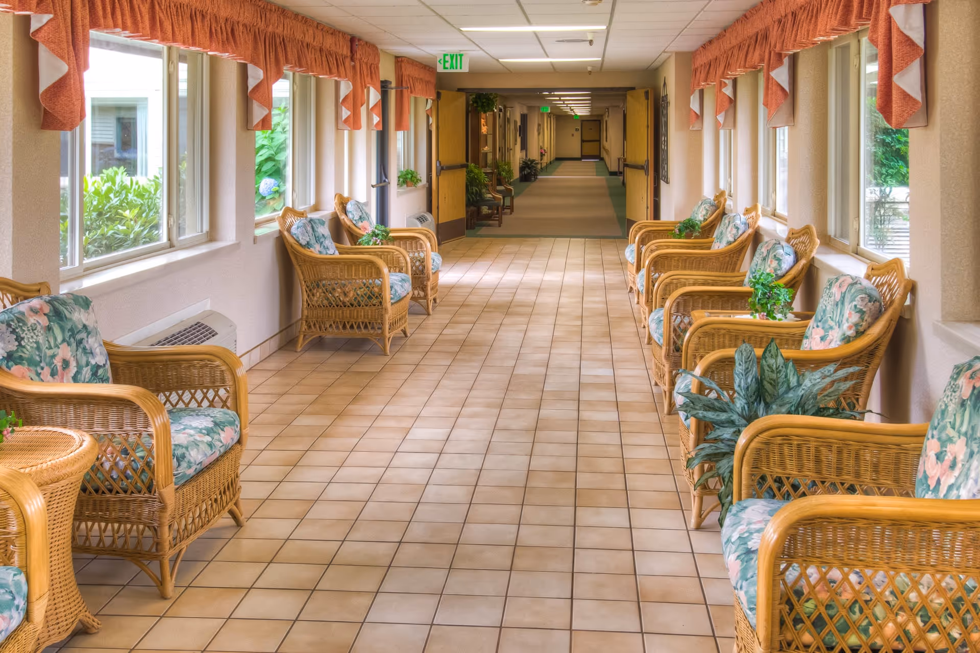 A bright hallway in a retirement community with large windows on one side, adorned with orange valance curtains. The hallway features wicker chairs with floral cushions and small potted plants on side tables. The floor is tiled, and the hallway extends into a carpeted corridor with more chairs and plants visible in the distance.
