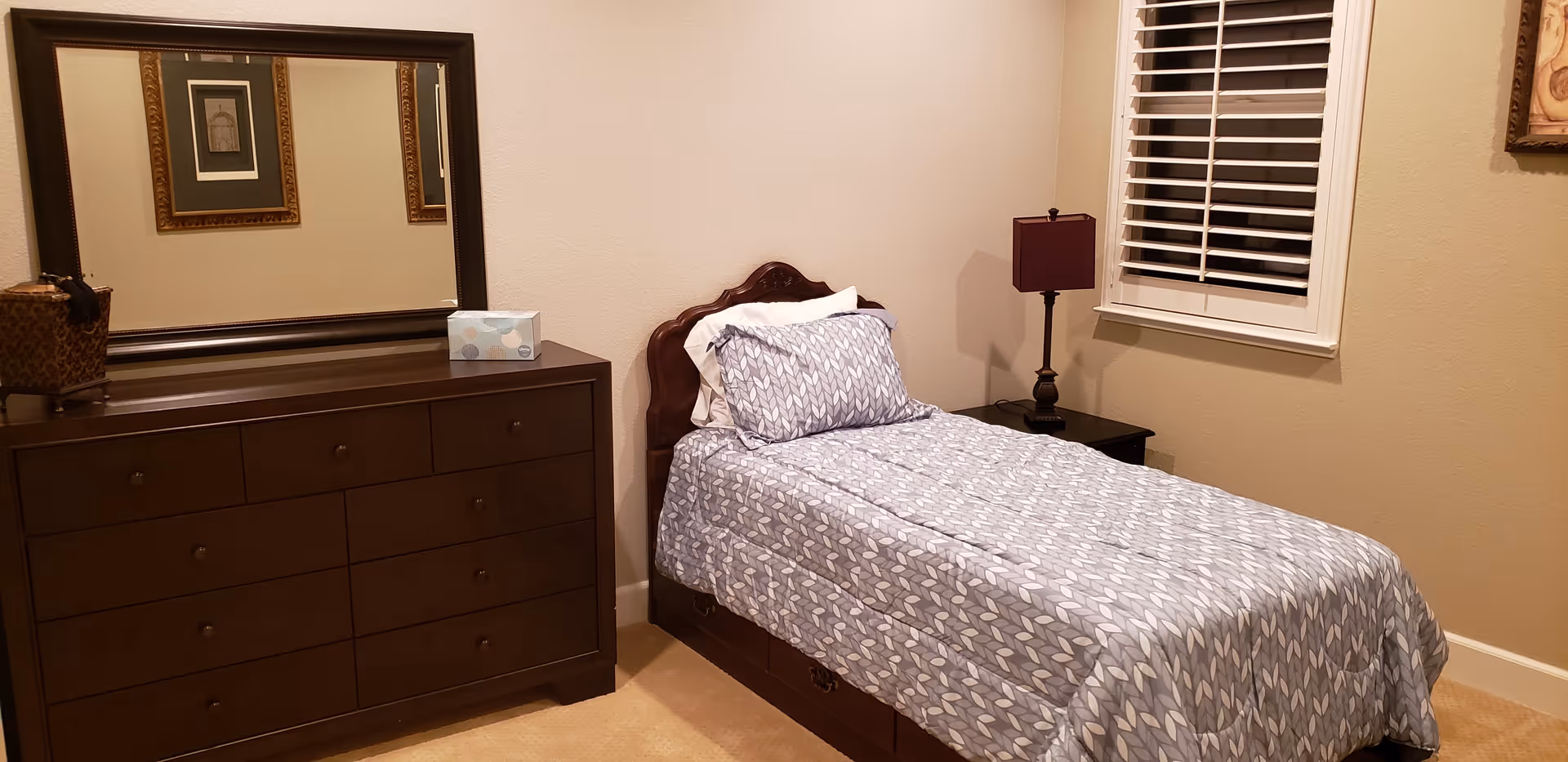 A neatly made single bed with patterned bedding beside a dark wood dresser, bedside table and lamp in a small bedroom.