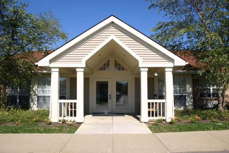 Front exterior view of a single-story building with a covered entrance supported by four white columns. The building has beige siding, large windows with white curtains, and is surrounded by green shrubs and trees under a clear blue sky.