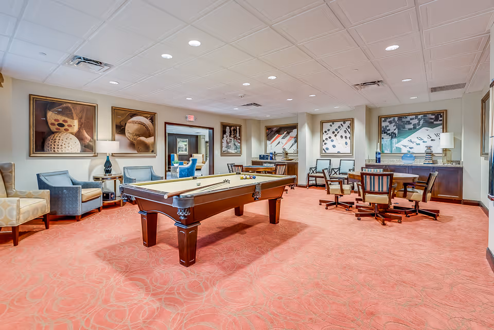 A spacious recreational room with a pool table in the center, surrounded by comfortable chairs and a round table with chairs on the right. The room has a red patterned carpet, beige walls adorned with framed artwork featuring sports themes, and a white ceiling with recessed lighting.
