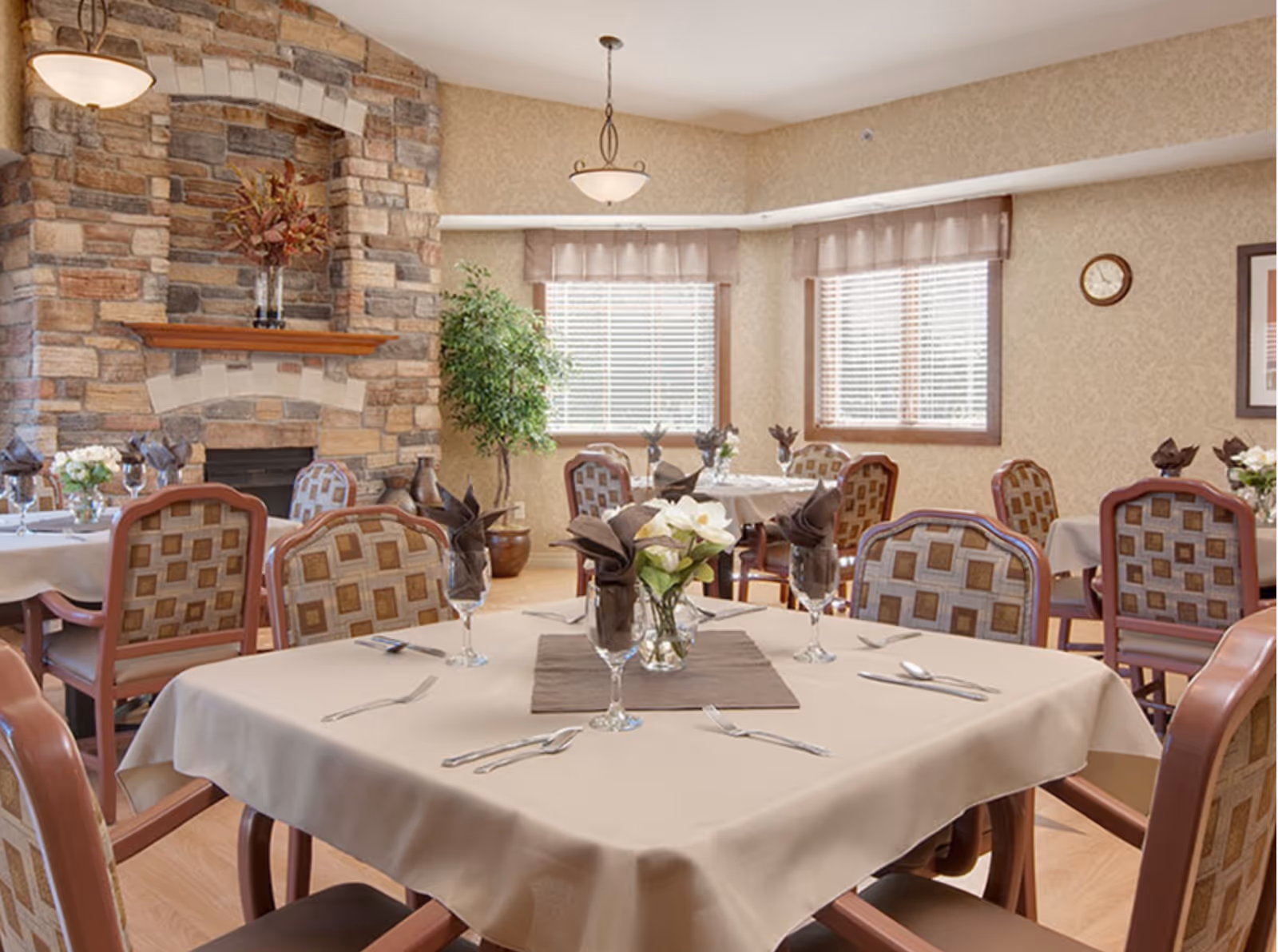 Bright dining room with neatly set tables, upholstered chairs, a stone fireplace, and large windows.