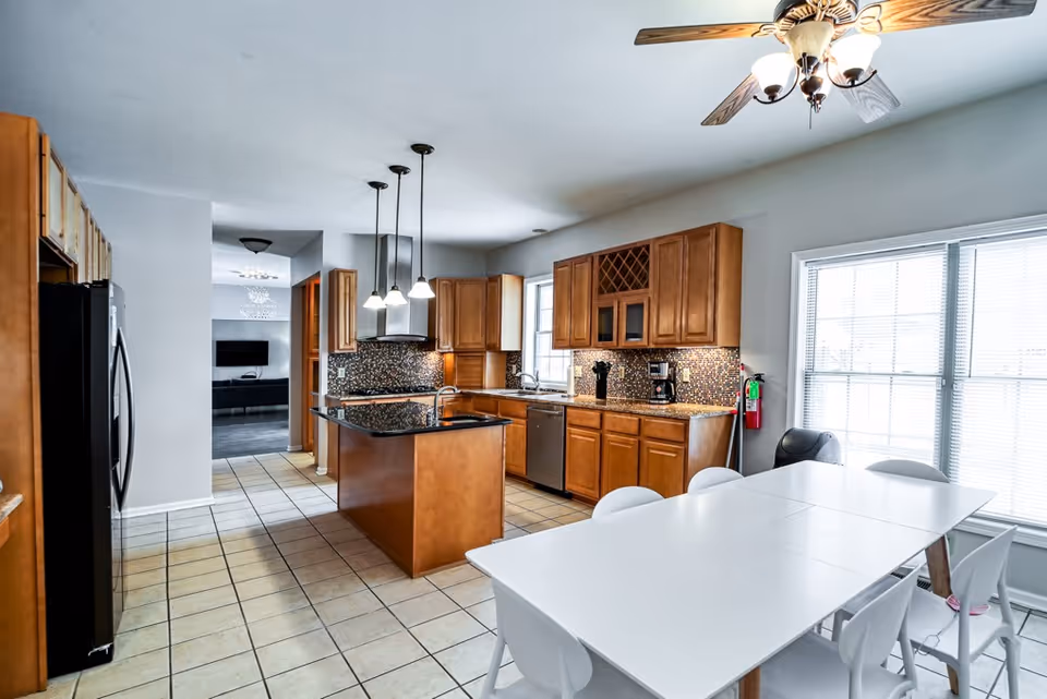 Bright kitchen with wooden cabinets, a central island with a black countertop, stainless steel appliances, and a white dining table with white chairs near a large window with blinds. Ceiling fan with lights is visible above the dining area.