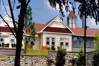 Exterior view of a single-story building with beige siding, white trim, and a red roof. The building features multiple windows and a small tower-like structure with a conical roof. The foreground includes a stone retaining wall, green shrubs, and trees partially framing the view.