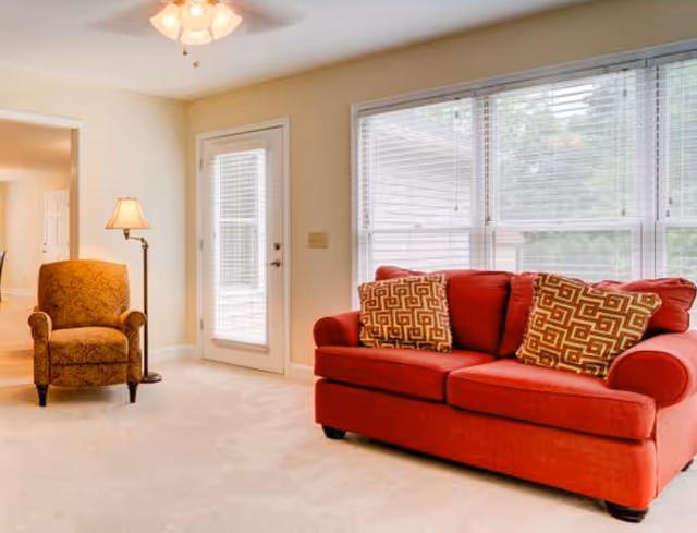 A bright living room with a red sofa adorned with two patterned cushions, a brown upholstered armchair, a floor lamp, and large windows with white blinds letting in natural light. There is a door with glass panels leading outside.