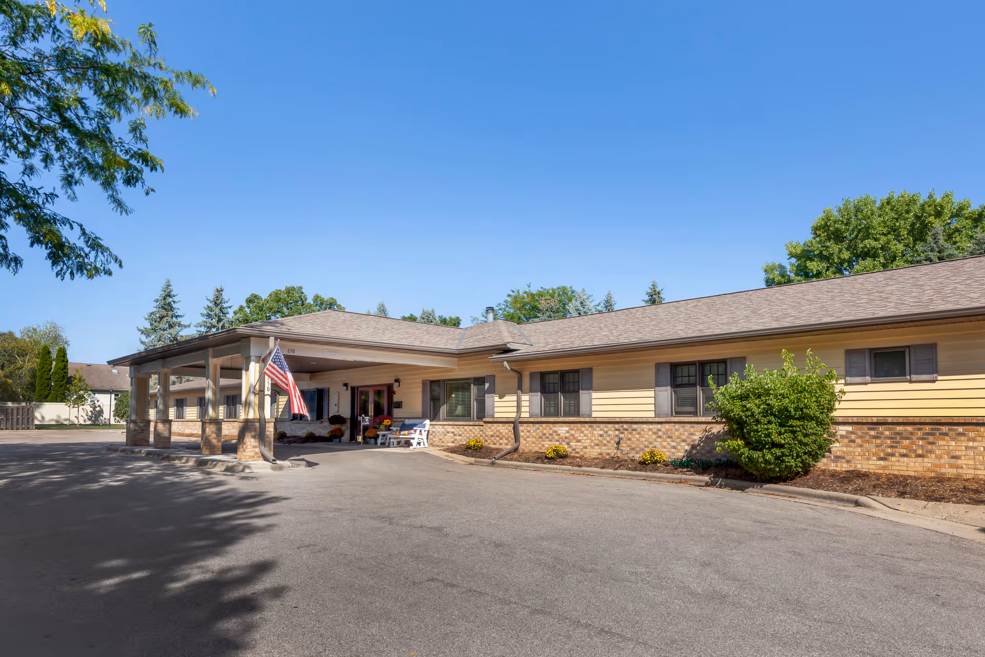 Exterior view of a single-story senior living facility building with a covered entrance, an American flag, and a bench near the entrance. The building has beige siding with brick accents and several windows. Trees and shrubs surround the building under a clear blue sky.