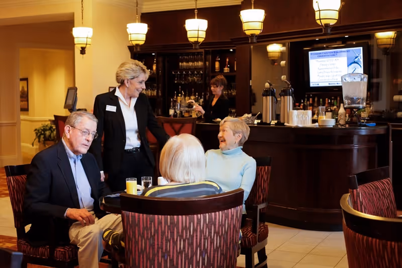 A group of three elderly people sitting around a table in a lounge area, engaged in conversation with a standing staff member. Behind them is a bar counter with a woman preparing drinks and various beverage containers and glasses on the counter.