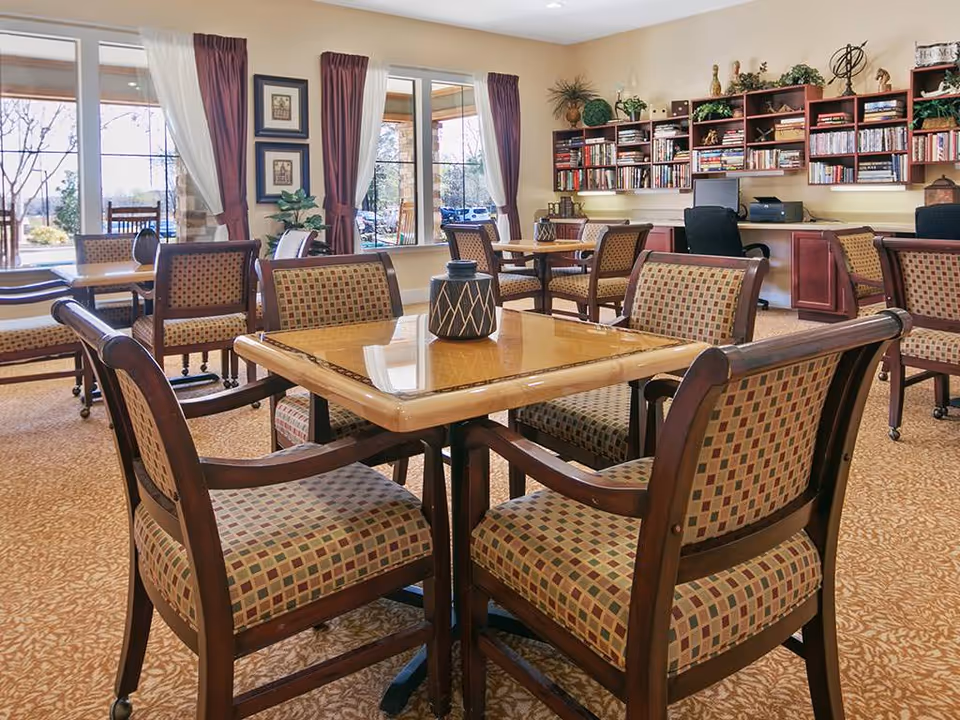 A well-lit common area with multiple wooden tables and cushioned chairs arranged neatly. The room features large windows with curtains allowing natural light to enter. There are bookshelves filled with books and decorative items along the back wall, and a computer workstation is visible. The carpet has a patterned design, and the overall atmosphere is warm and inviting.