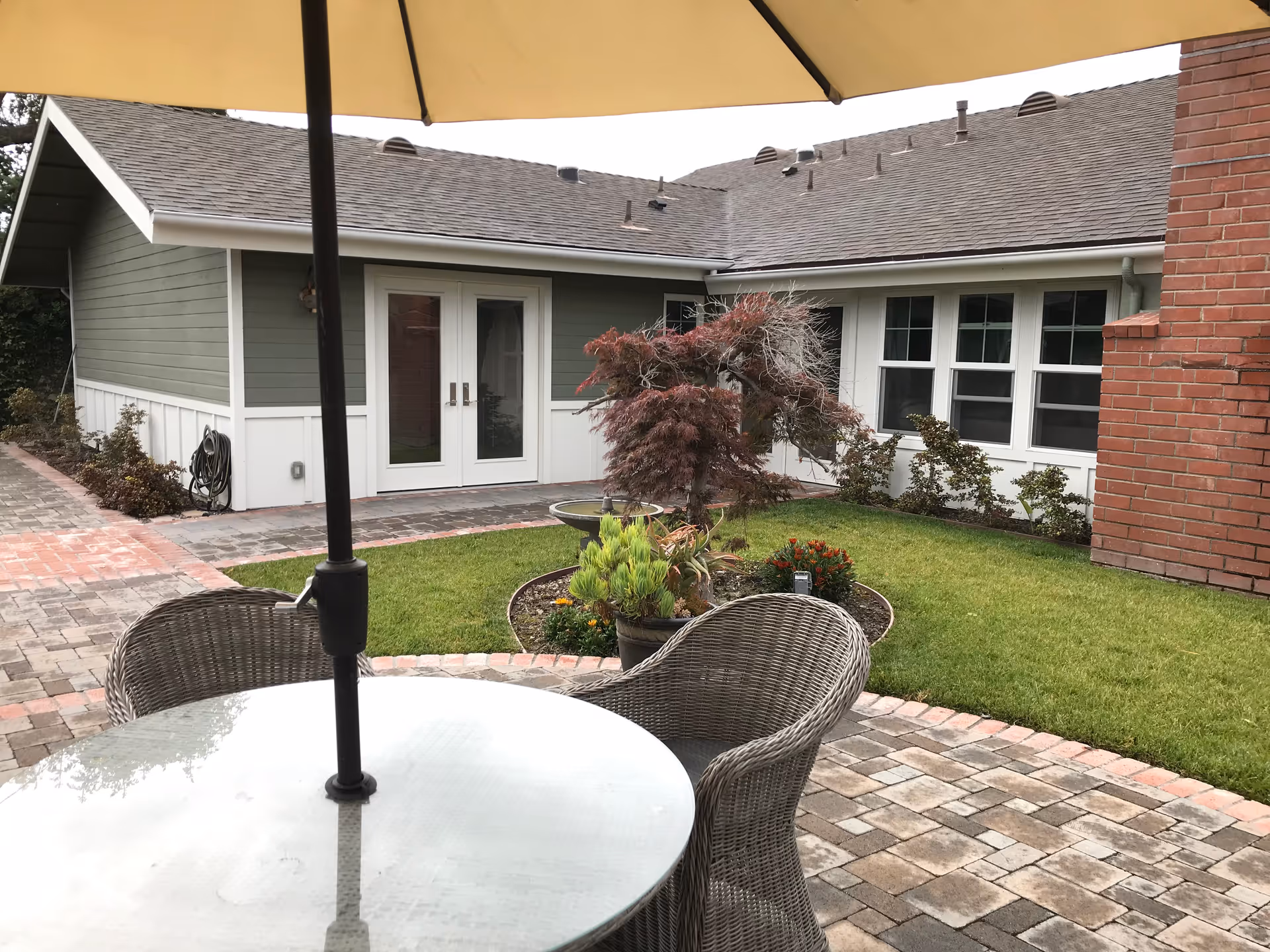 Outdoor patio area with a round glass table and wicker chairs under a large beige umbrella. The patio is paved with bricks and surrounded by a small garden with green grass, plants, and a small tree. In the background, there is a building with green and white siding, white-framed windows, and a brick chimney.