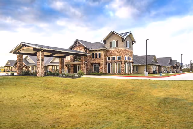 Exterior view of Discovery Village Clear Lake, showing a large building with stone and beige siding, multiple windows, and a covered entrance. The building is surrounded by a grassy area and a paved driveway under a partly cloudy sky.