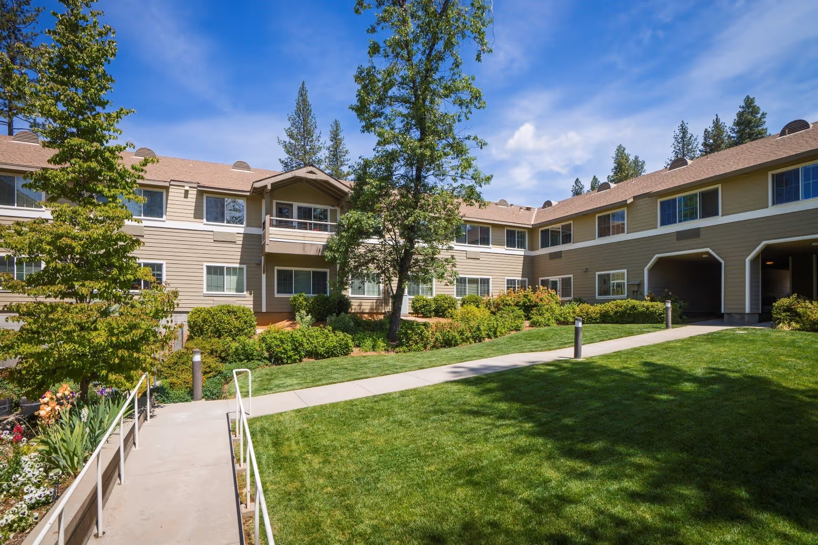Exterior view of Eskaton Village Grass Valley showing a two-story building with beige siding and multiple windows. The building is surrounded by well-maintained green lawns, bushes, trees, and a clear blue sky with some clouds. A concrete walkway with handrails leads through the landscaped area.
