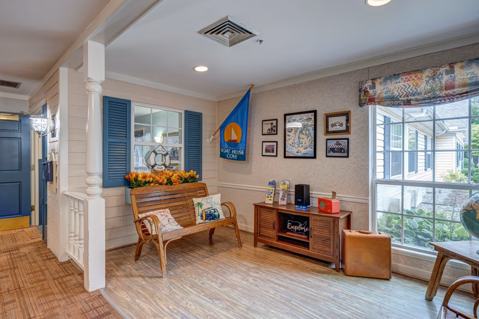 A cozy interior corner of a senior living facility featuring a wooden bench with decorative pillows and a planter of orange flowers. The wall behind the bench has blue window shutters and a life preserver decoration. Adjacent wall displays framed pictures and a blue flag with a sailboat and the words 'Boat House Cove.' Below the pictures is a wooden cabinet with brochures and a small speaker. A large window with patterned valance lets in natural light, showing greenery outside. The floor is wood laminate, and the hallway to the left has a blue door and carpeted floor.