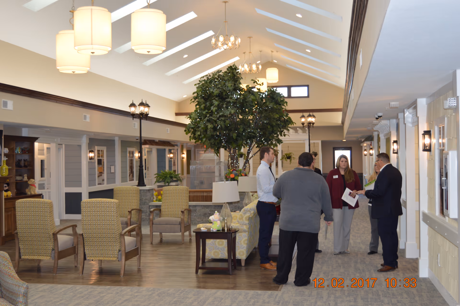 A bright communal lounge in an assisted living facility with seating, indoor plants, chandeliers and a small group of people talking.