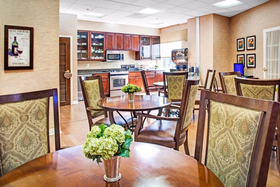 A bright dining area with round wooden tables and upholstered chairs featuring a green patterned fabric. Each table has a small vase with green flowers. In the background, there is a kitchen with wooden cabinets, a microwave, stove, and a coffee machine near a window. The walls are decorated with framed pictures and a wine country poster.