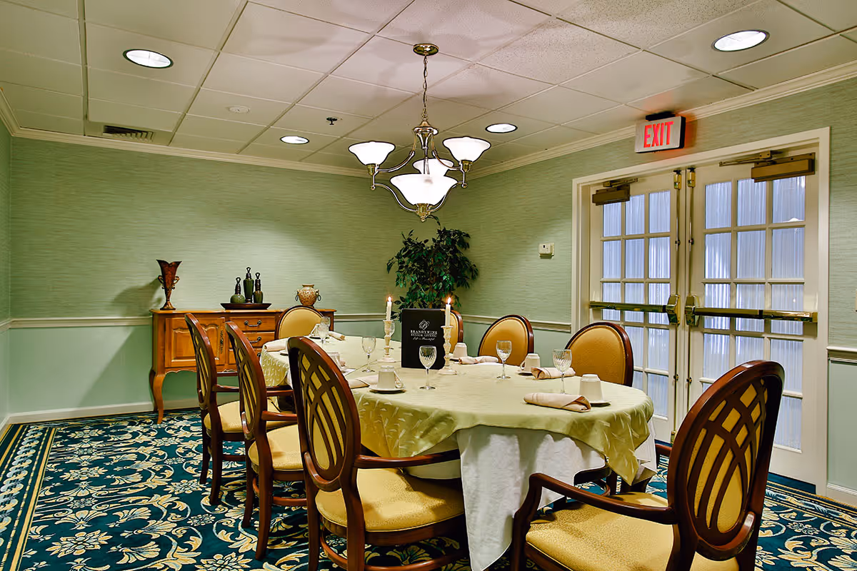 A formal dining room with a long oval table covered with a light green tablecloth and set with glassware, napkins, and candles. The room has six upholstered chairs with wooden frames around the table, a sideboard with decorative items against the wall, a green plant in the corner, patterned carpet, and double glass doors with an exit sign above them.