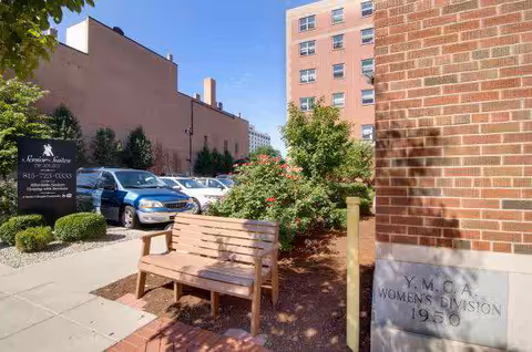 Outdoor area of a senior living facility with a wooden bench, a garden with bushes and flowers, parked cars, and a brick building with a plaque that reads 'Y.M.C.A. Women's Division 1950'. A sign for Senior Suites is visible near the parked cars.
