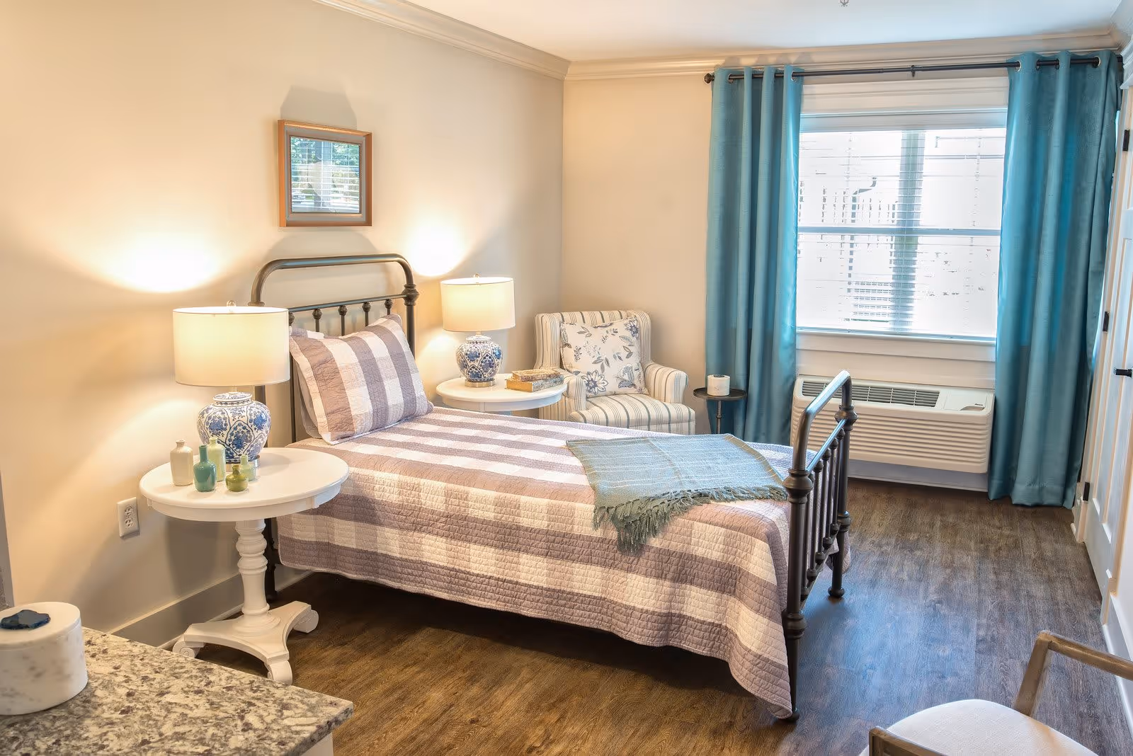 A cozy bedroom with a single bed featuring a checkered quilt and pillow. There are two bedside tables with matching blue and white lamps, a small armchair with floral and striped upholstery, a window with teal curtains, and a wooden floor.