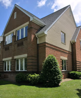 Brick two-story house-like building with windows, shrubs, and a green lawn under a blue sky.