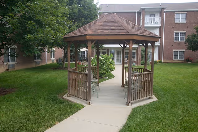 Wooden gazebo with benches on a paved path in a grassy courtyard in front of a brick senior living building.