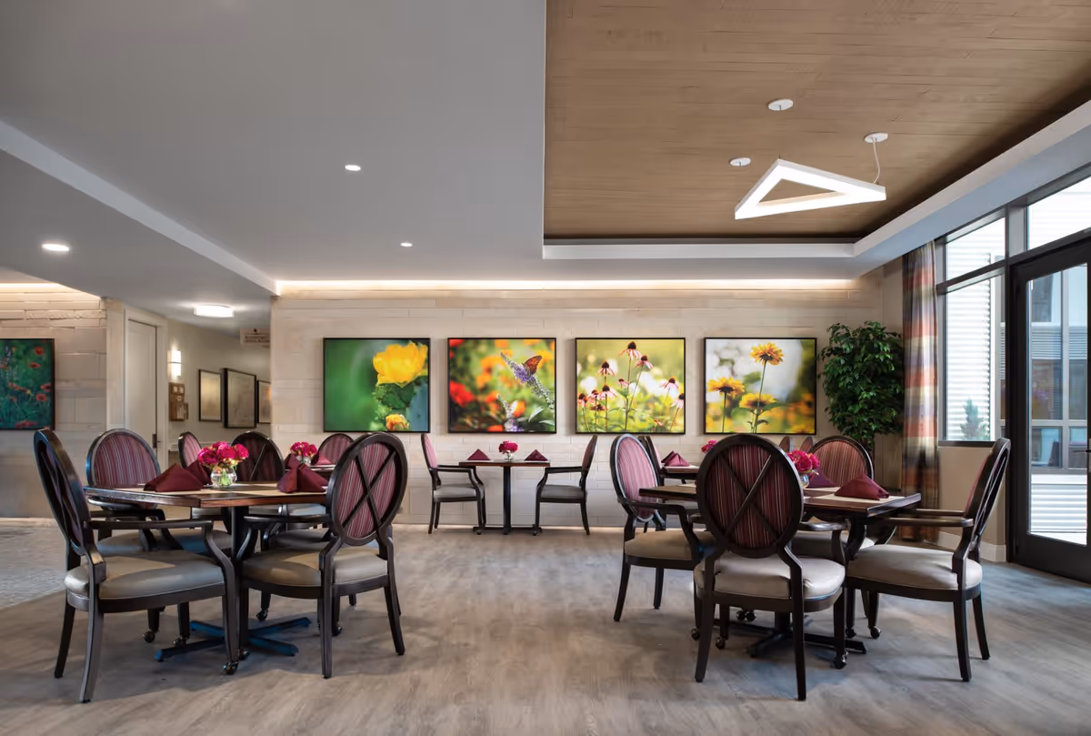 Bright dining room with round tables and upholstered chairs, colorful floral artwork on the wall, and large windows.