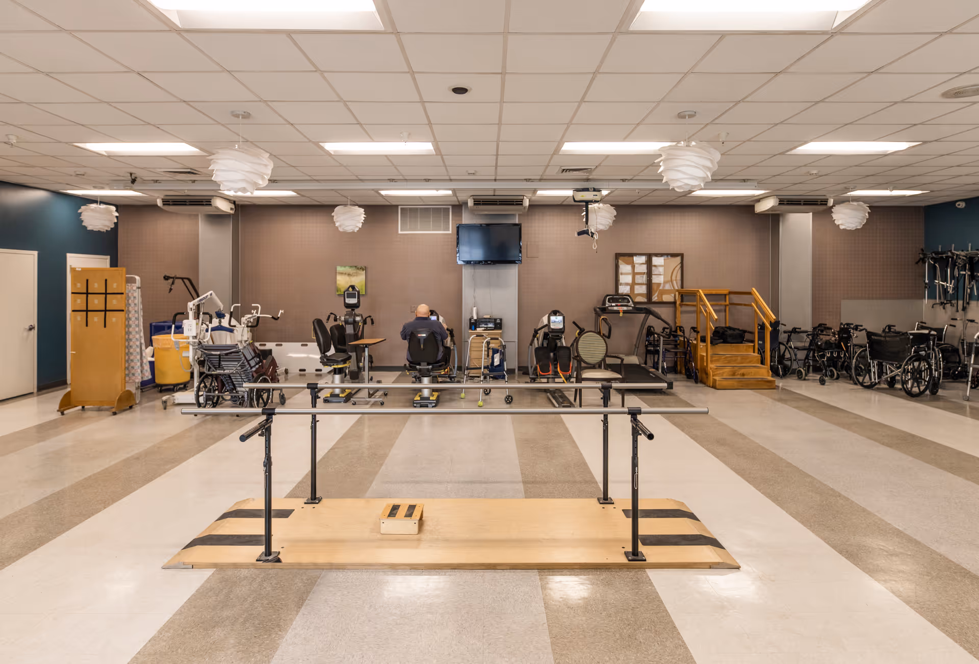 A spacious rehabilitation or physical therapy room with exercise equipment including parallel bars, stationary bikes, treadmills, and various mobility aids such as wheelchairs and walkers. The room has a tiled floor, ceiling lights, and a TV mounted on the wall.