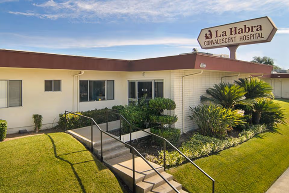 Front entrance of La Habra Convalescent Hospital with a large sign, walkway, and landscaped lawn.