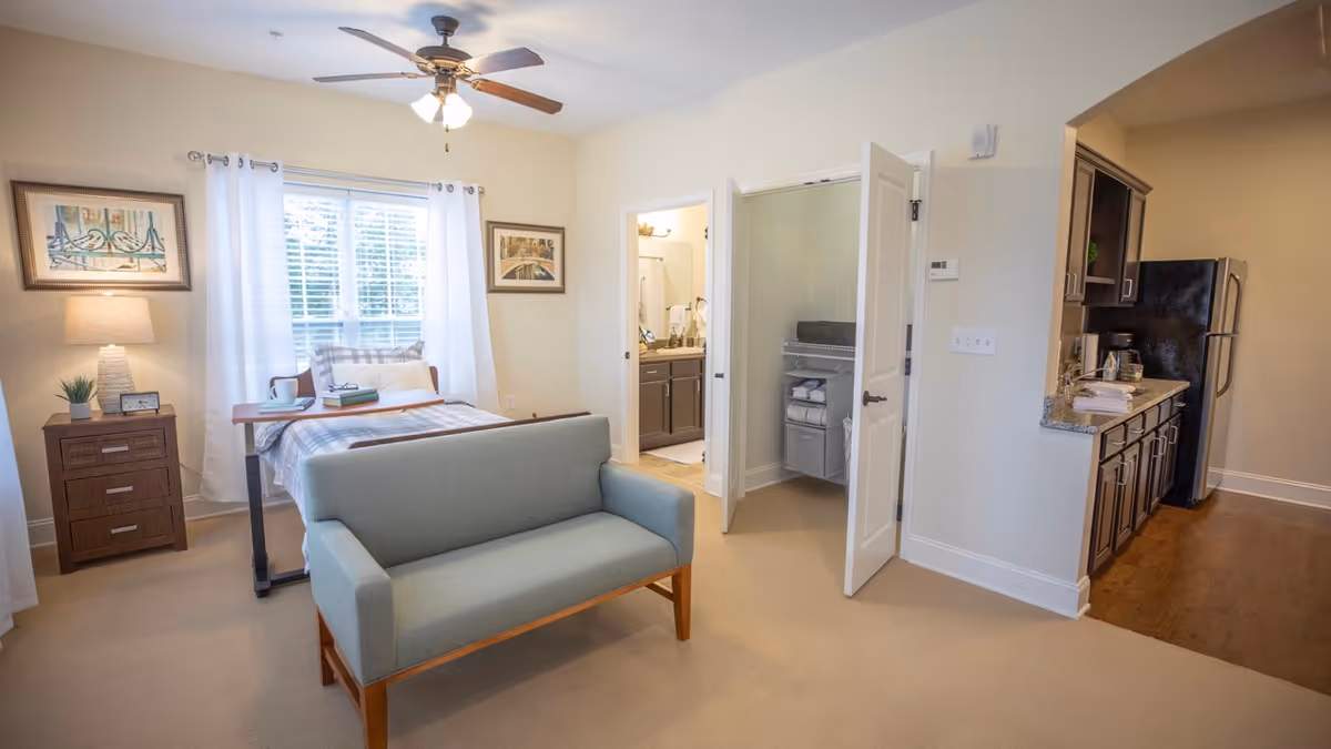 A well-lit senior living facility room featuring a bed with a tray table, a small blue loveseat, a nightstand with a lamp and plant, two framed pictures on the walls, an open door revealing a bathroom, and a kitchenette area with dark cabinets and a refrigerator.