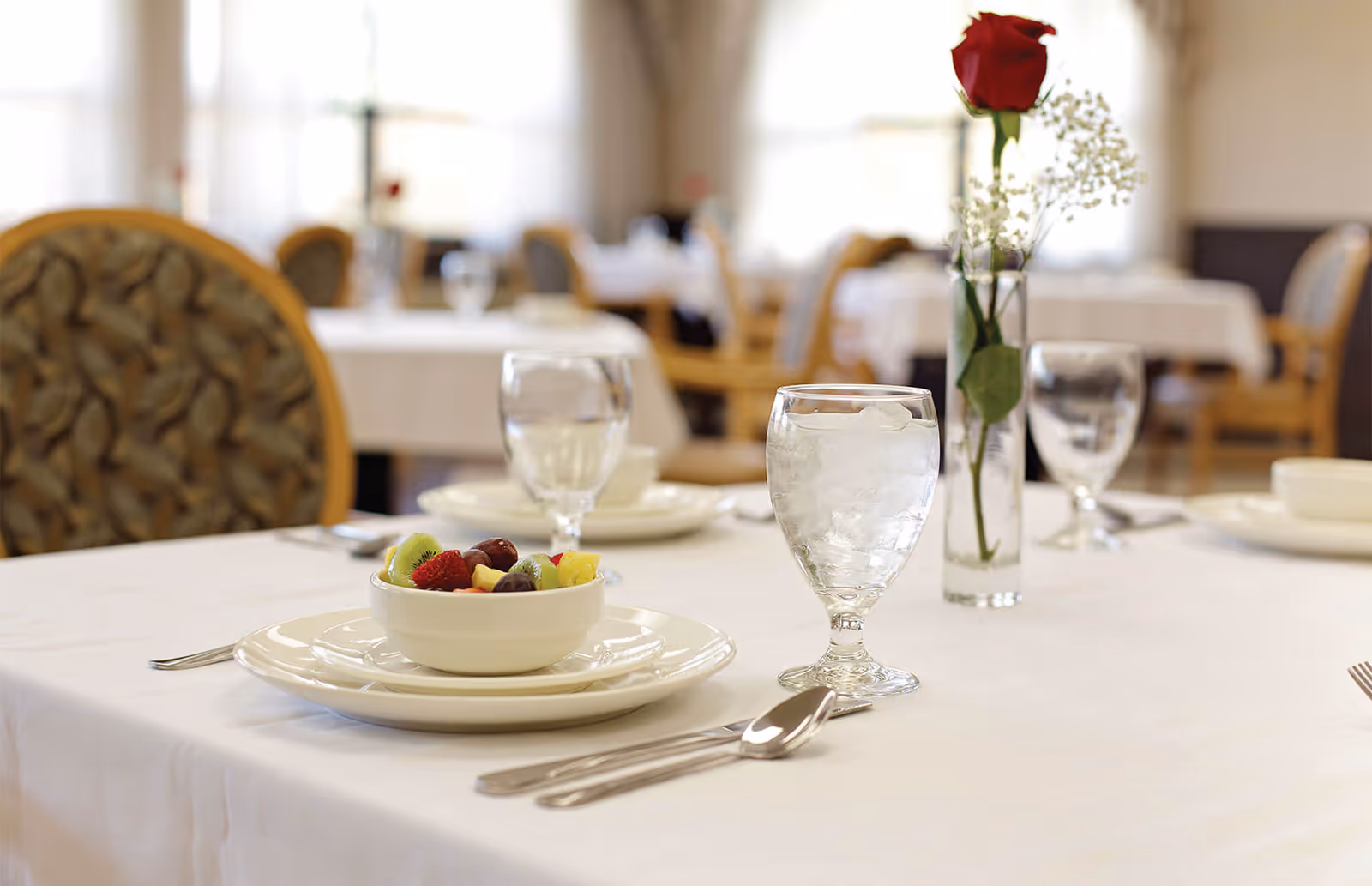 A neatly set dining table with a bowl of fruit, water glasses, silverware, and a single red rose in a vase in a bright dining room.