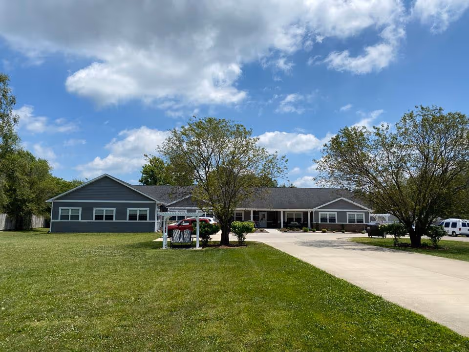 A single-story building with gray siding and a dark roof, surrounded by green grass and trees under a partly cloudy blue sky. A concrete driveway leads up to the building entrance, with a few parked vehicles visible on the right side.