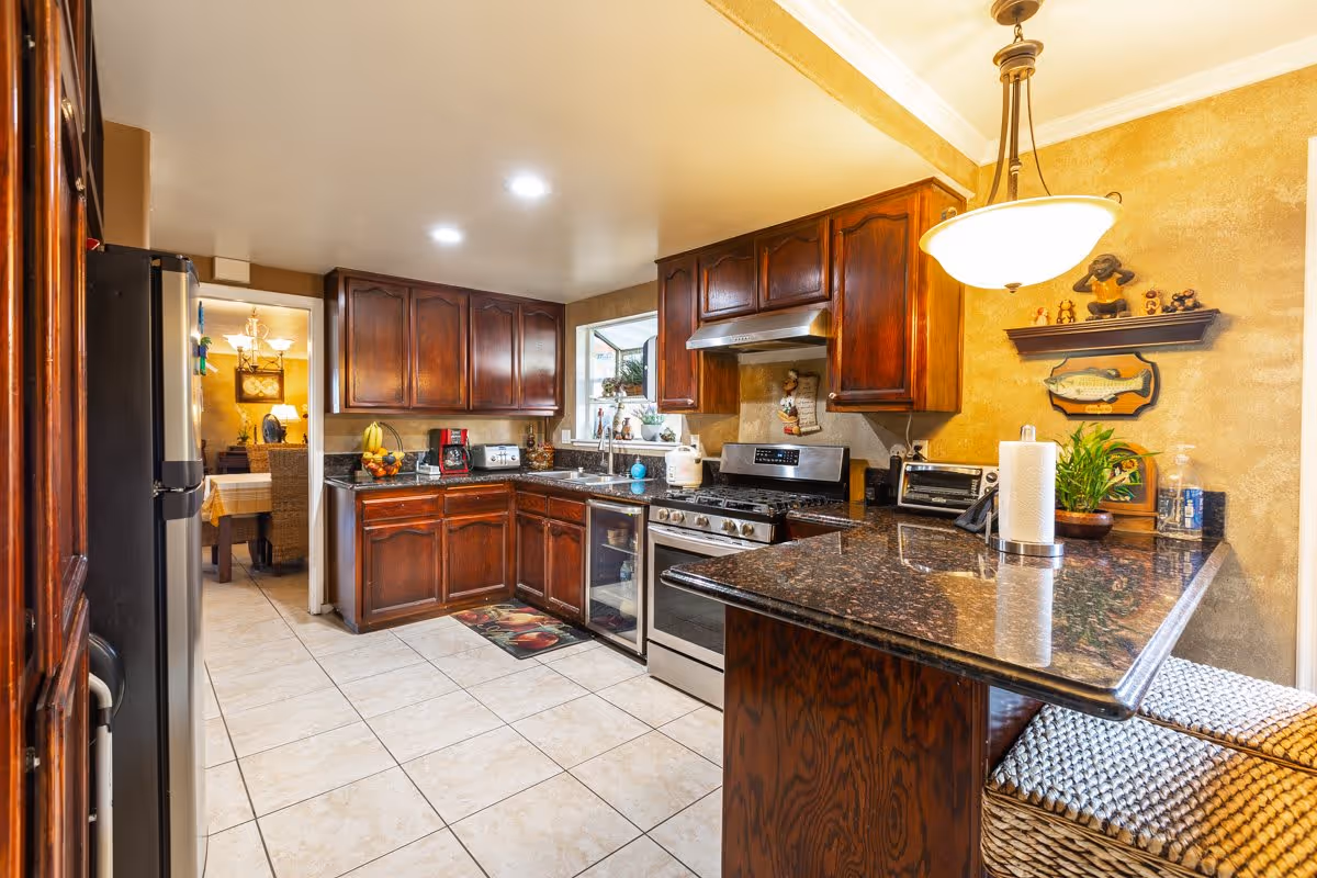 A well-lit kitchen with dark wooden cabinets, granite countertops, and stainless steel appliances including a stove and refrigerator. There is a window above the sink, various kitchen appliances on the counters, a hanging light fixture, and a small shelf with decorative items on the wall. The floor is tiled and a dining room is visible through a doorway.