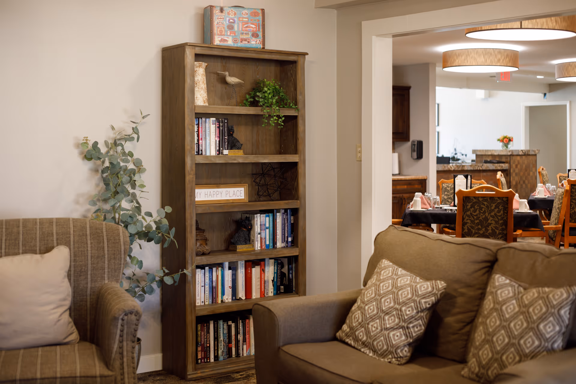 Cozy living room area with a brown armchair and a brown sofa with patterned cushions. A wooden bookshelf filled with books and decorative items stands against a beige wall. In the background, a dining area with tables and chairs is visible, set with napkins and cups, under round ceiling lights.