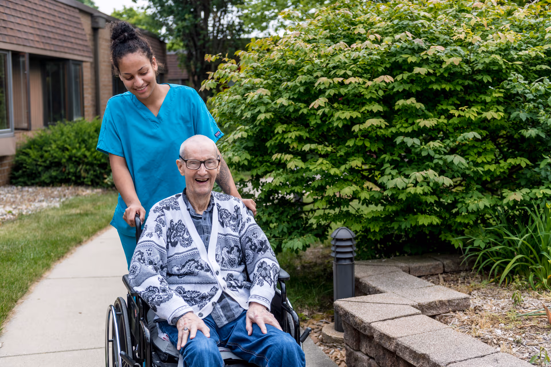A smiling elderly man in a wheelchair is being pushed along a sidewalk by a smiling caregiver wearing blue scrubs. They are outside near a building with green bushes and plants in the background.