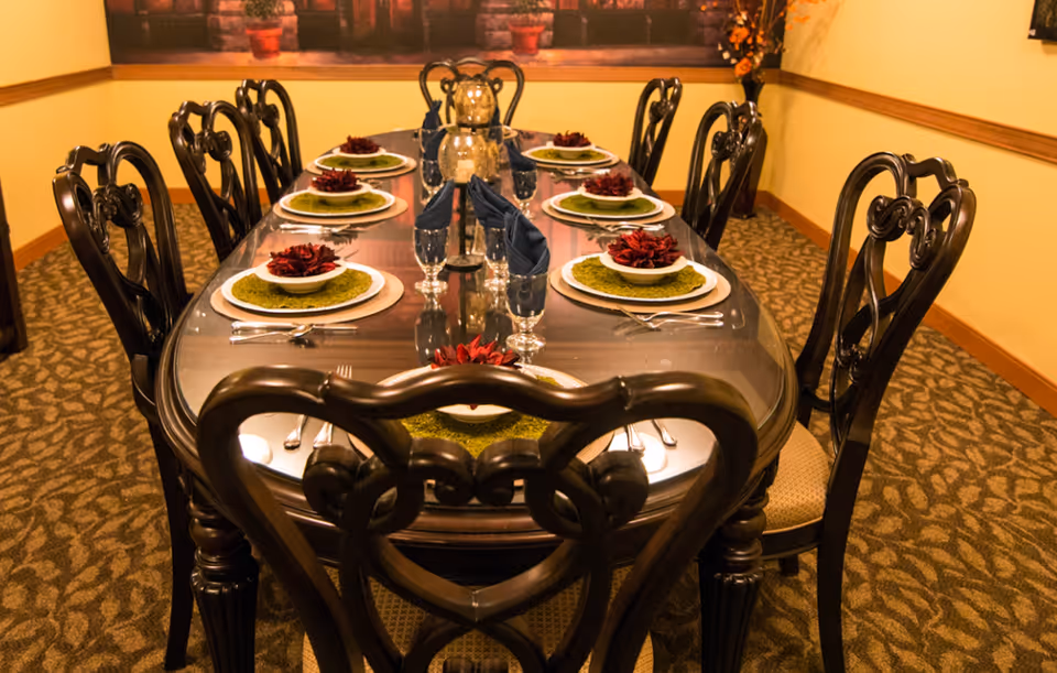 A formal dining room with a long wooden table set for eight people. Each place setting includes a white plate with a green placemat and a decorative red flower on top, silverware, and a glass with a folded navy blue napkin. The room has patterned carpet, yellow walls with wooden trim, and a large framed picture on the wall.