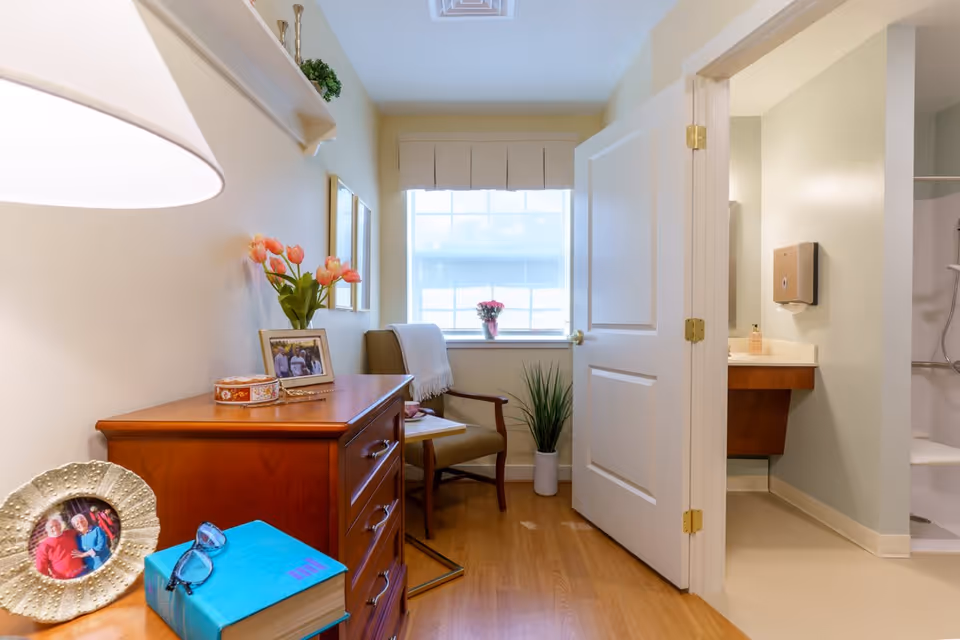 A cozy senior living room area with a wooden dresser topped with framed photos, a blue book with glasses, and a vase of pink tulips. A comfortable armchair with a white throw blanket is positioned near a window with a small potted plant on the windowsill. An open door reveals a bathroom with a sink, soap dispenser, and a shower area.